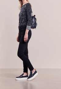 Top nero a maniche lunghe con motivo floreale bianco, jeans skinny neri e sneakers slip-on nere con suola bianca e accento rosso nel tallone.