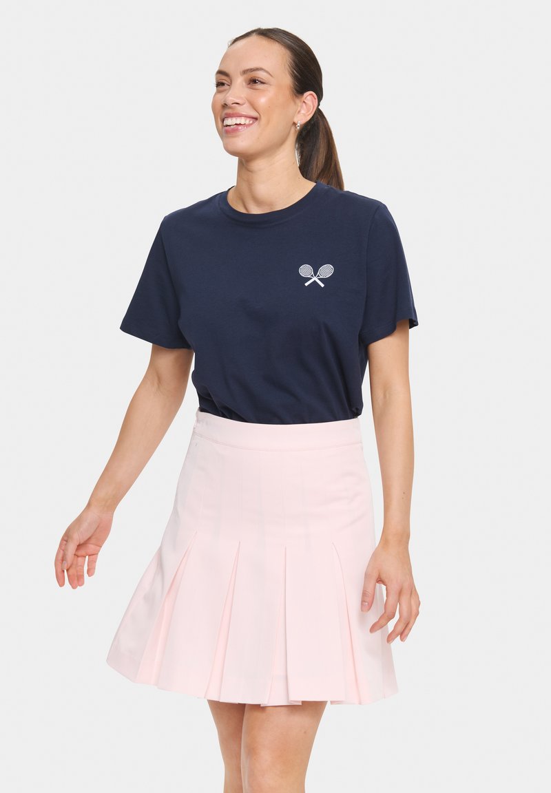 Navy cotton t-shirt with a white crossed racquet logo, paired with a pink pleated skirt. The outfit features a relaxed fit and soft texture.
