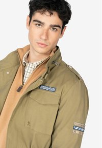 Chaqueta verde oliva con cremallera, dos bolsillos en el pecho, un parche en el lado izquierdo del pecho y una capa interna en tono beige, llevada sobre una camisa de cuadros.