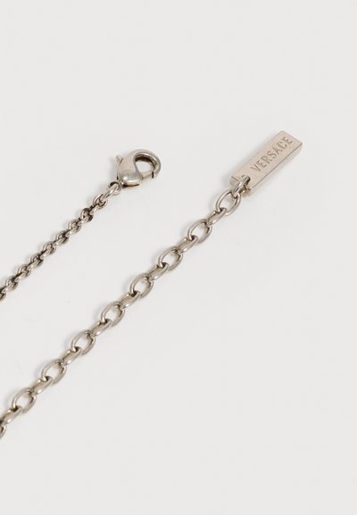 Collier chaîne en argent avec fermoir mousqueton et plaque rectangulaire gravée "VERSACE" sur fond blanc.
