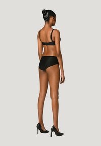 Svart bh och högmidjade trosor av mjukt material, med justerbara axelremmar. Modellen bär svarta högklackade skor och poserar från baksidan.