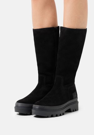Černé semišové kozačky střední výšky s robustními gumovými podrážkami a bočními zipy, nošené na bosých nohou na jednobarevném bílém pozadí.