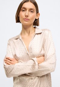 Bluză de culoare bej deschis, realizată dintr-un material mătăsos, cu un model geometric, mâneci lungi, guler și închidere cu nasturi.