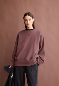 Junge Frau mit glattem braunem Haar trägt einen oversized mauve Sweatshirt und schwarze Hosen, während sie ein dunkel strukturiertes Kleidungsstück vor einem mauve Hintergrund hält.