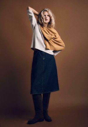 Traper suknja u A-liniji s visokim strukom, uparena s kremastim puloverom i šalom smiješane boje senfa prebačenim preko ramena. Smeđe čizme do koljena.