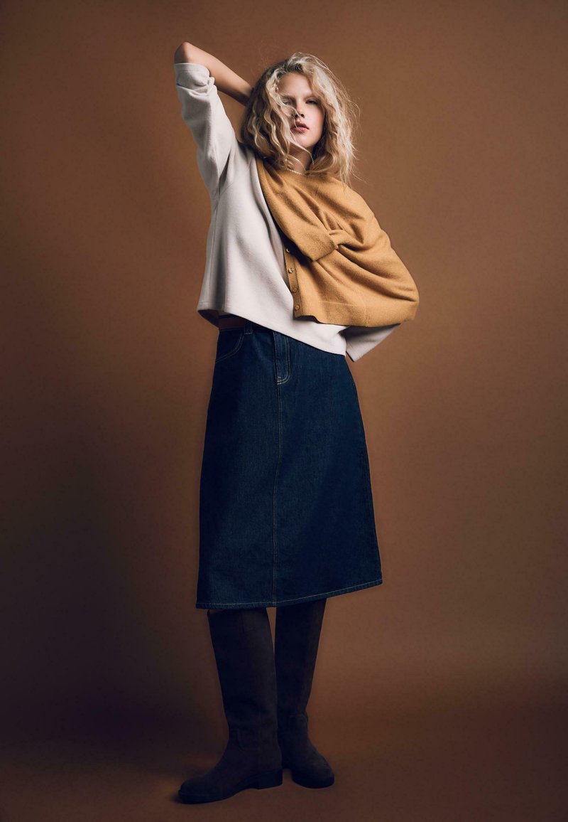 Dżinsowa spódnica w kształcie litery A z wysokim stanem, połączona z kremowym swetrem i musztardowym szalem dzianinowym narzuconym na ramiona. Brązowe kozaki do kolan.