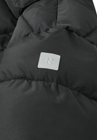 Schwarzer gepolsterter Jackenstoff mit einem kleinen quadratischen reflektierenden Patch, auf dem ein stilisiertes "R"-Logo aufgenäht ist.
