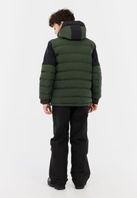 Groene pufferjack met een zwarte capuchon en schouderaccenten, voorzien van horizontale gestikte secties; gecombineerd met zwarte broeken en laarzen.
