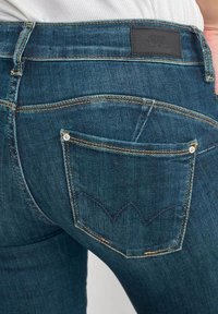 Donkerblauwe denim jeans met een getailleerd ontwerp, voorzien van een achterzak met een decoratief stikselpatroon en een leren logo patch op de tailleband.