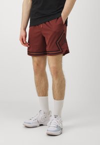Pantalones cortos deportivos rojos con acentos negros, textura de malla y logo lateral. Combinados con zapatillas blancas y calcetines de caña media.