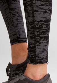 Czarno-szare legginsy w wzór z teksturowanym designem, o dopasowanym kroju i szwach szytych, w parze z ciemnymi butami sportowymi.