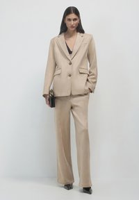 Donna che indossa un completo in velluto beige con top nero, mentre tiene una pochette nera, in piedi su uno sfondo grigio chiaro uniforme.