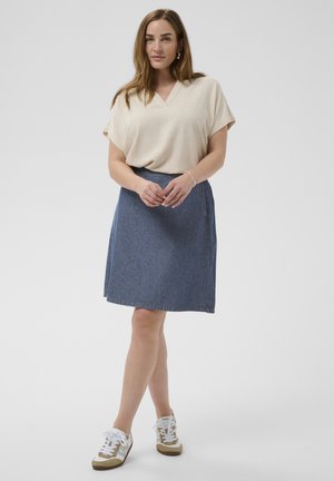 Kvinde stående iført beige kortærmet V-hals top, blå denim knælængde nederdel og hvide sneakers på en ensfarvet baggrund.