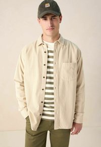 Beige corduroy shirt met knopen, over een gestreepte olijf- en crème T-shirt. Olijfgroene broek en een donkergroene pet met een patchdetail.