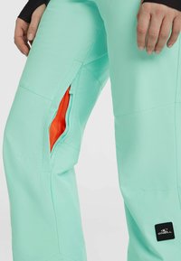 Mintgroene sneeuwbroeken hebben ritszakken met oranje mesh voering. Het materiaal is duurzaam met zichtbare naden en een logopatch.