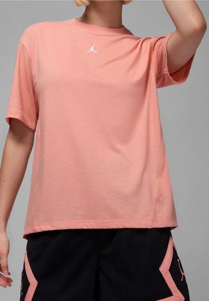 Person iført en kortærmet fersken-farvet Jordan t-shirt og sorte shorts med fersken-farvet Jordan-logo, der løfter en arm mod en ensfarvet grå baggrund.