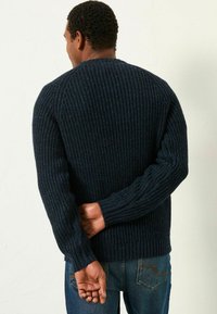 Mann mit dunkler Hautfarbe, der einen marineblauen, gerippten Strickpullover und blaue Jeans trägt, und mit hinter dem Rücken verschränkten Händen steht.