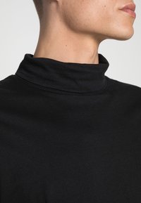 Czarny golf wykonany z gładkiego materiału. Posiada wysoki kołnierz i prosty design, z widocznymi szwami wzdłuż krawędzi.