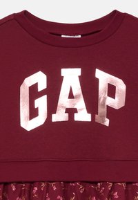 Bordo dukserica s natpisom "GAP" u metalik srebrnim slovima, u kombinaciji s suknjom s cvjetnim uzorkom; mekan materijal, dizajn s okruglim izrezom.