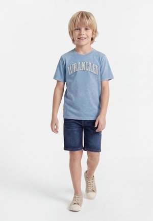 Blondýnský chlapec se usmívá, kráčí vpřed, má na sobě modré tričko Wrangler, tmavé džínové šortky a béžové tenisky na bílém pozadí.