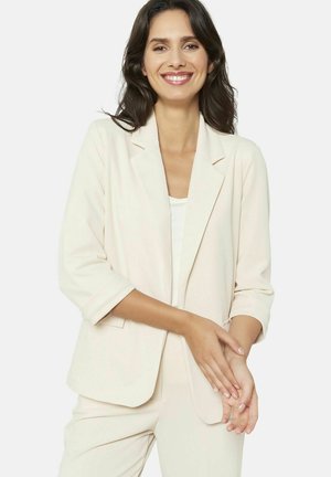 Heledad helebeez blazer, millel on vabalt voolav lõige, klassikaline krae, kolmeveerandik sleeves ja pehme tekstuur. Kandmise all on valge topp.