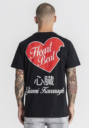 Mężczyzna z tatuażami na ramionach, ubrany w czarną koszulkę z dużym czerwonym sercem i napisem „Heart Beat”, znakami chińskimi oraz „Gianni Kavanagh” na plecach.