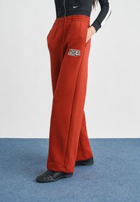 Κόκκινο παντελόνι φόρμας Nike Club, κατασκευασμένο από μείγμα βαμβακιού, με ελαστική μέση, πλαϊνές τσέπες και κέντημα με λογότυπο. Σχεδιασμός με φαρδιά γραμμή στο πόδι.