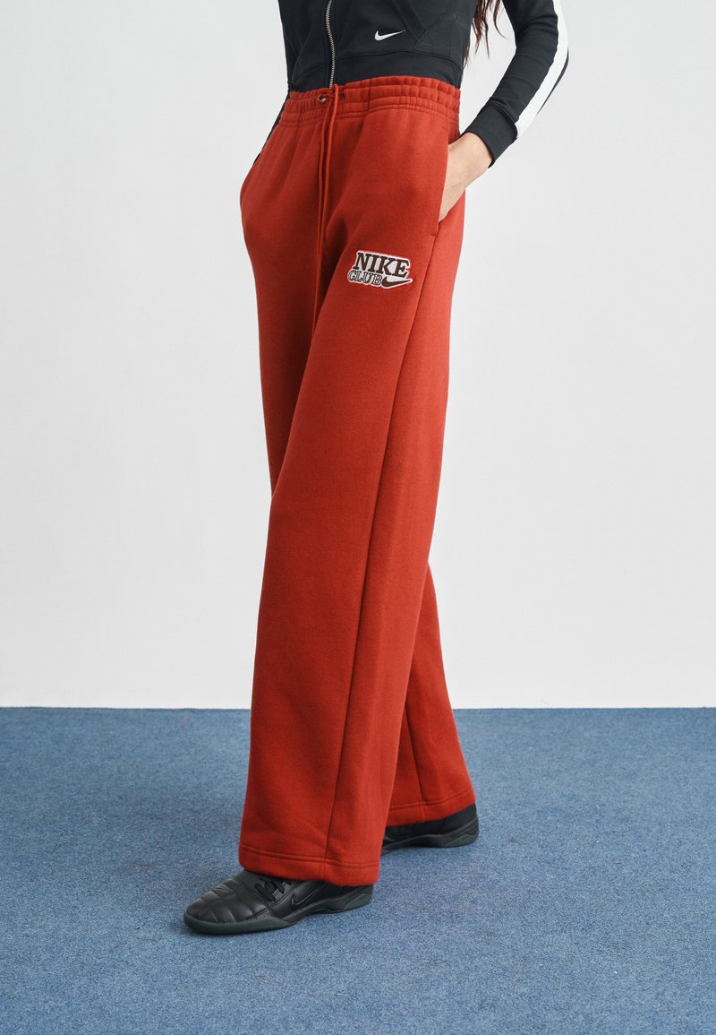 Κόκκινο παντελόνι φόρμας Nike Club, κατασκευασμένο από μείγμα βαμβακιού, με ελαστική μέση, πλαϊνές τσέπες και κέντημα με λογότυπο. Σχεδιασμός με φαρδιά γραμμή στο πόδι.