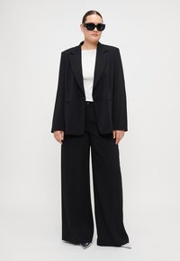 Črn prilagojen blazer z zarezanimi reverji, v kombinaciji z belim topom in širokimi hlačami; model nosi prevelike črne sončne očala.