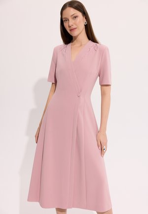 Kobieta ubrana w jasnoróżową sukienkę kopertową z krótkim rękawem i dekoracyjnymi detalami na ramionach, stojąca na jednolitym tle.