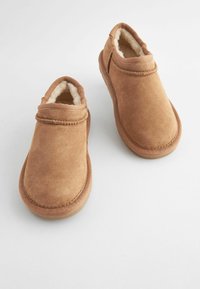 Scarpe slip-on in suede marrone con un morbido rivestimento color crema, punta rotonda e dettagli cuciti. Suola in gomma piatta con una finitura testurizzata.