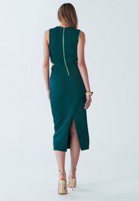 tealfärgad midiklänning med dragkedja bak, sidoutskärningar, elegant silhuett och slits bak. Kombinerad med guldiga sandaletter med remmar.
