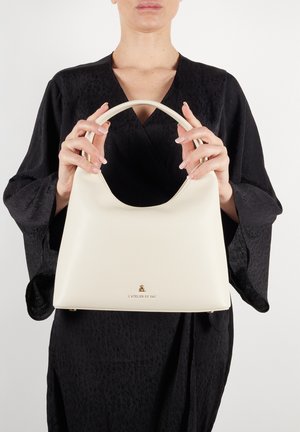 Kremowa skórzana torebka z zakrzywionym uchwytem, o gładkiej fakturze i złotym logo na dole.