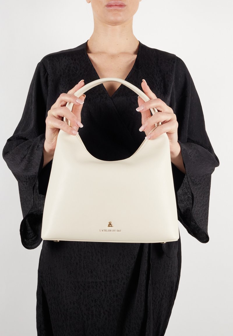 Kremowa skórzana torebka z zakrzywionym uchwytem, o gładkiej fakturze i złotym logo na dole.