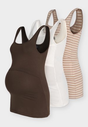 Tehotná žena v béžovom pruhovanom tielku a čiernych krátkych nohaviciach, držiaca veľkú čiernu kabelku, stojaca na jednoliatom pozadí.