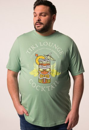 Hombre que lleva una camiseta verde con un diseño colorido de cóctel tiki y el texto "Tiki Lounge Cocktail" mirando hacia un lado con una mano en el bolsillo.