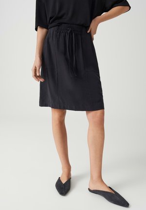 Femme portant une jupe noire mi-longue avec des poches avant et des chaussures noires à bout pointu sans lacets, debout devant un fond clair.