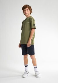 Camiseta de algodón verde oliva con mangas cortas, logo en el lado izquierdo del pecho, combinada con pantalones cortos azul marino y zapatillas blancas con detalles en negro.