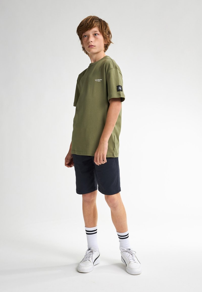 Camiseta de algodón verde oliva con mangas cortas, logo en el lado izquierdo del pecho, combinada con pantalones cortos azul marino y zapatillas blancas con detalles en negro.