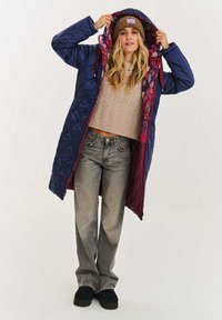 Pikowany długi płaszcz w ciemnym niebieskim kolorze z kwiatową podszewką; posiada kaptur, przedni suwak z regulowanym sznurkiem oraz luźny krój. Beżowy, strukturalny sweter.