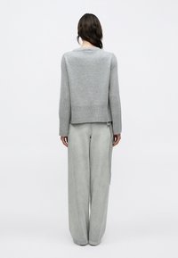 Femme aux cheveux foncés portant un pull gris clair et un jean gris clair à jambes larges, debout de dos sur un fond blanc uni.