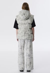 Lichtgrijze puffervest met een kap, voorzien van zwarte tekst en patronen. Gecombineerd met grijze broek met wijde pijpen die vergelijkbare ontwerpen tonen.