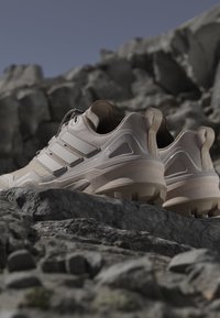 Buty wędrowne Terrex z lekką beżową syntetyczną cholewką, trzema poziomymi paskami, teksturowanymi panelami i solidną podeszwą z bieżnikiem.