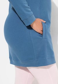 Robe sweat bleue avec des manches longues et une poche, accompagnée de leggings rose clair. Texture en coton lisse, design décontracté.