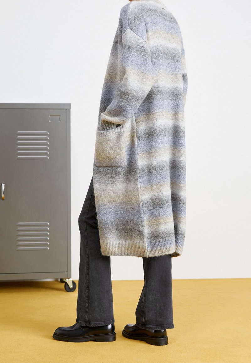Personne portant un long gilet rayé doux avec des poches, un pantalon noir et des chaussures noires, debout sur un sol jaune près d'un meuble métallique gris.