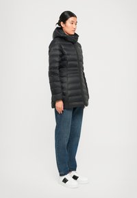 Zwarte pufferjas met een capuchon, voorzien van een ritsluiting, horizontaal doorgestikt patroon en geribde manchetten, gecombineerd met blauwe jeans en witte sneakers.