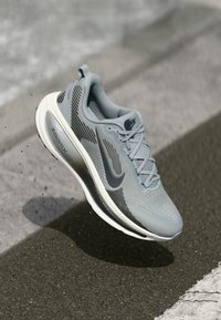 Γκρι αθλητικό παπούτσι με υφασμάτινη σόλα και υφή, μαύρες λεπτομέρειες και μια εξέχουσα λογότυπο "Zoom" στο πλάι, με καμπύλο σχέδιο σόλας.