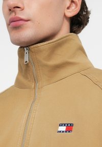 Μπεζ ζακέτα με φερμουάρ, φτιαγμένη από ύφασμα με λεία υφή. Διαθέτει ψηλό γιακά και ένα μικρό κεντημένο λογότυπο σε κόκκινα, άσπρα και μπλε χρώματα.