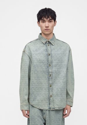 Jeune homme portant une chemise en jean bleu clair à motifs avec des boutons marron, debout devant un fond blanc uni.