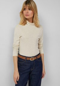 Beiger Langarm-Rollkragenpullover mit weicher Textur, kombiniert mit dunkelblauen Jeans und einem braunen Gürtel mit einer runden silbernen Schnalle.